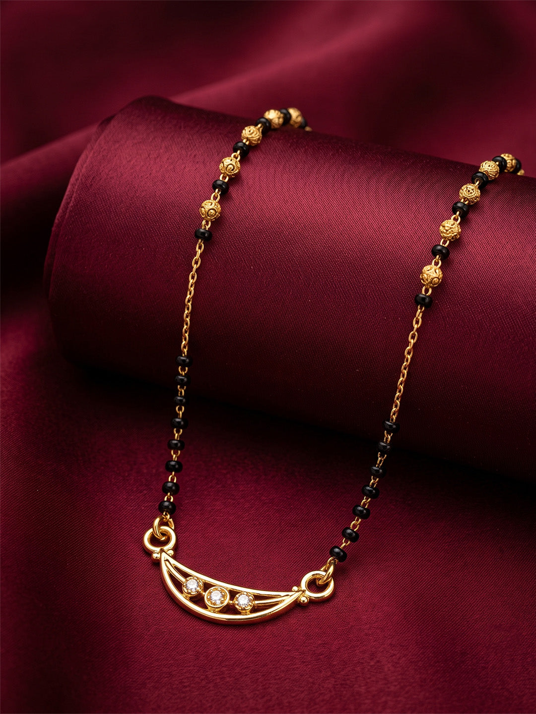 A gold-toned necklace with a crescent moon pendant set with small clear stones, accented by black and gold beads, displayed on a dark red satin fabric background lfariya jewellery.